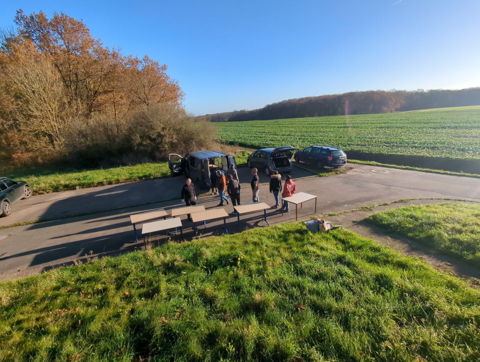 Vue sur un parking avec plusieurs voitures ou camionettes et des bénévoles autour avant d'y charger des tables.